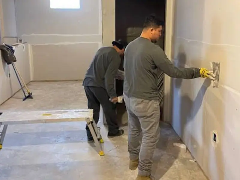 Two men applying plaster or drywall compound to interior walls in a room under construction. Fixed by a experienced restoration company in San Diego